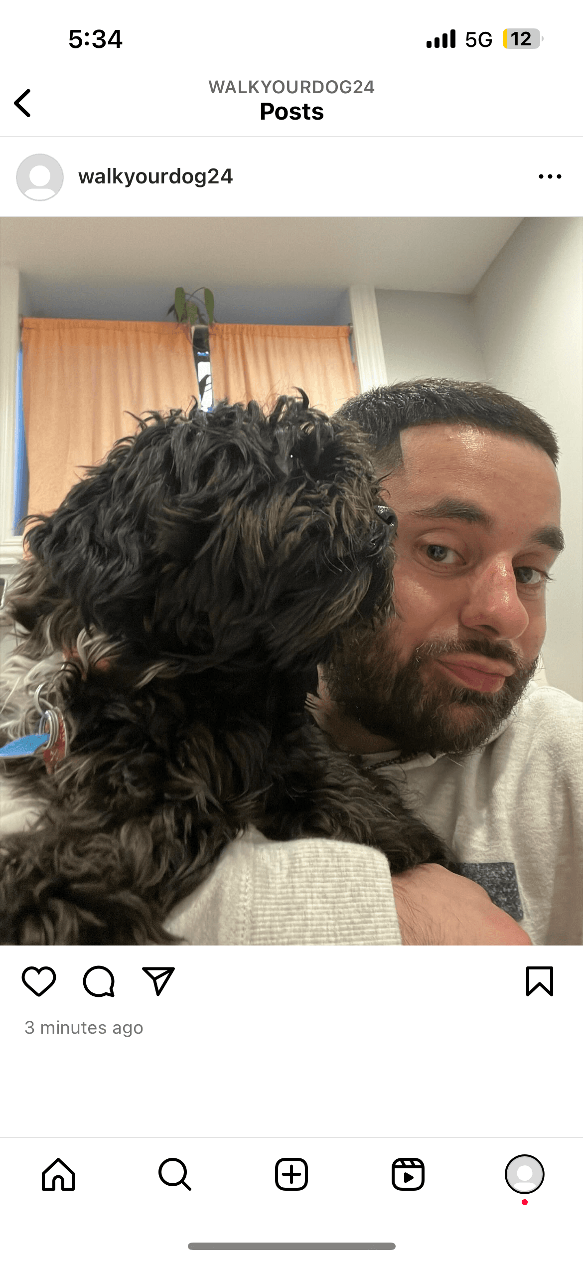 A bearded man cuddles a small, curly-haired dark dog against his shoulder indoors.