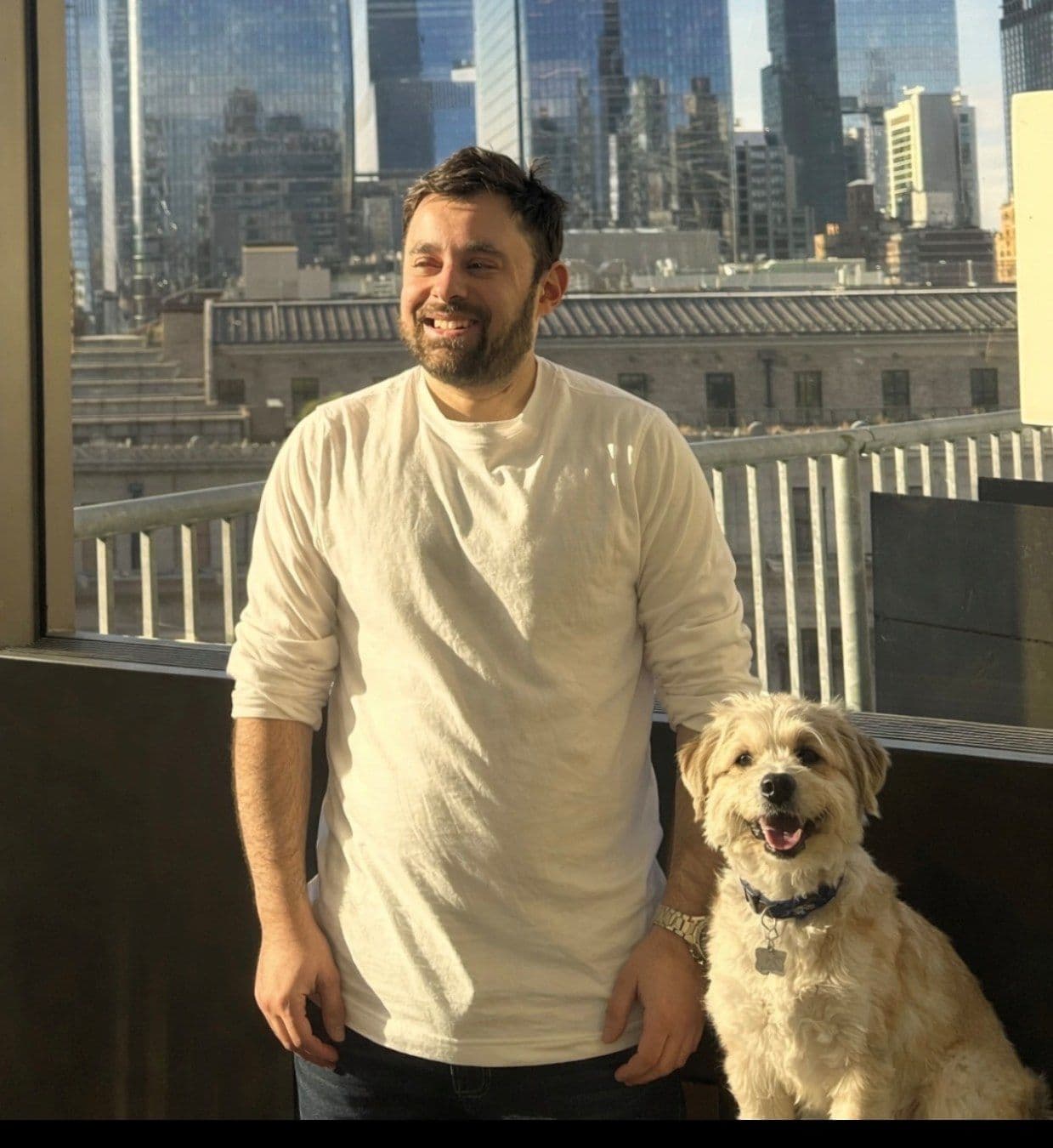 A smiling man and his small dog stand on a balcony overlooking a city skyline.